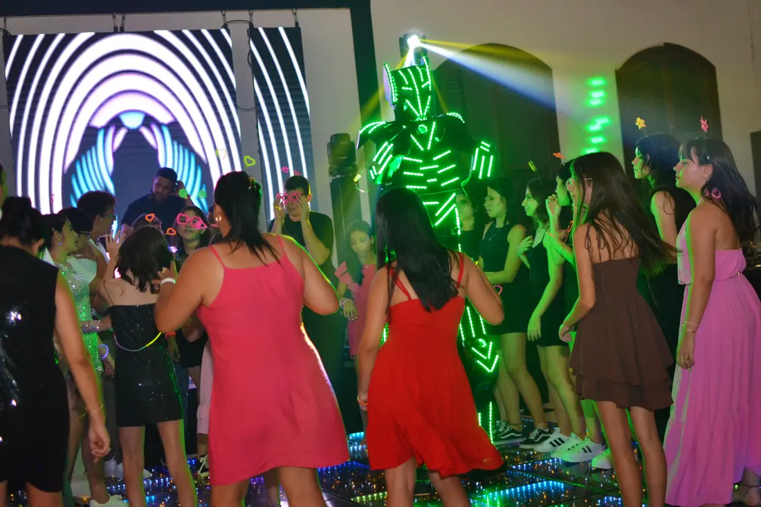 Convidados na pista de uma festa de 15 anos, com iluminação colorida e uma atração com traje de LED.