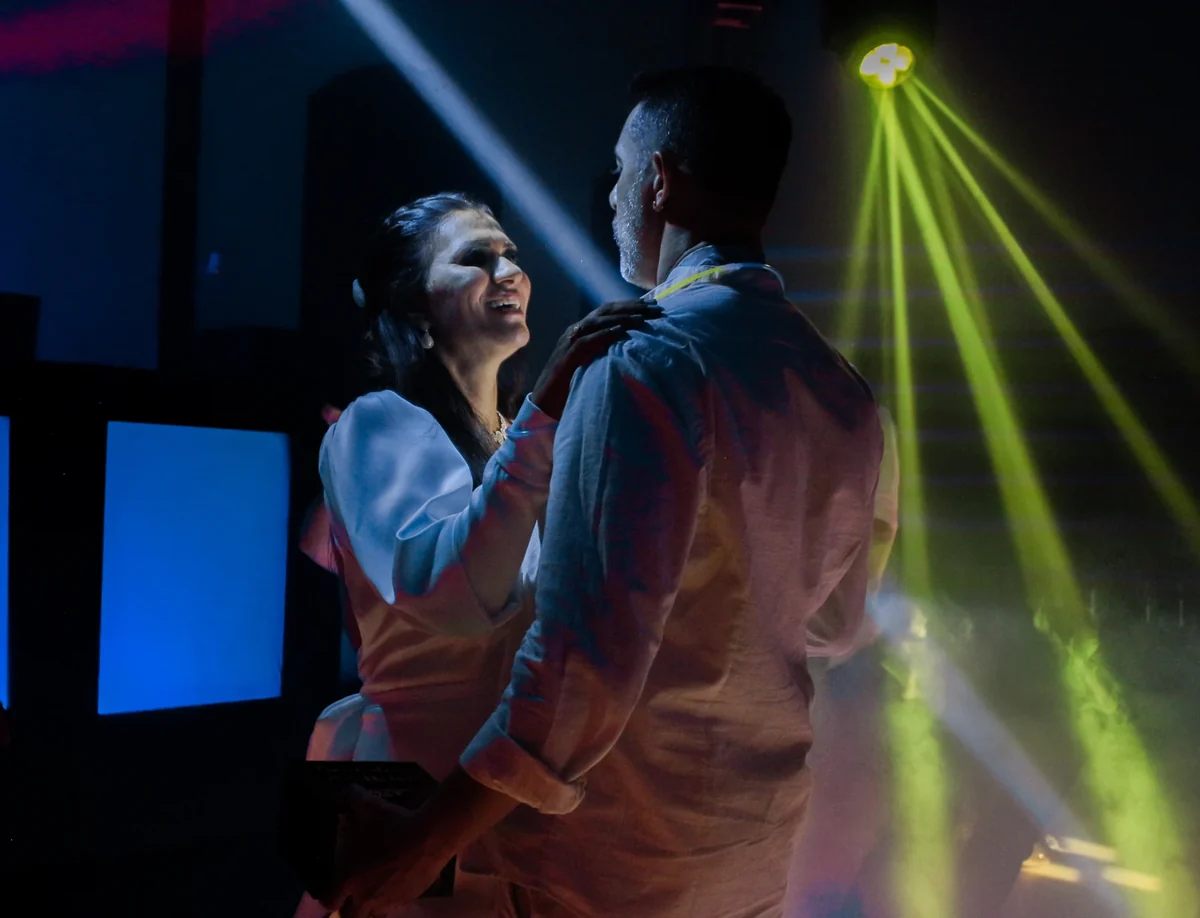 Um casal dança de mãos dadas e sorri sob as luzes amarelas e azuis da pista.