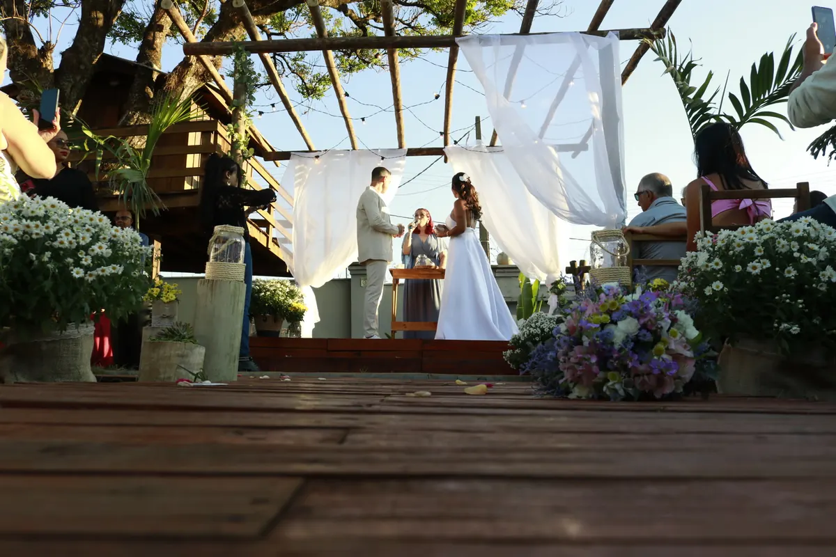 Casal diante da celebrante em cerimônia ao ar livre, com flores e cortinas brancas.