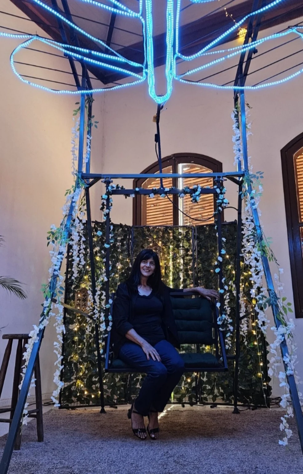 Dona Adélia, sócia fundadora da Ki Festa Eventos, sentada em um balanço decorado com flores e iluminação azul.