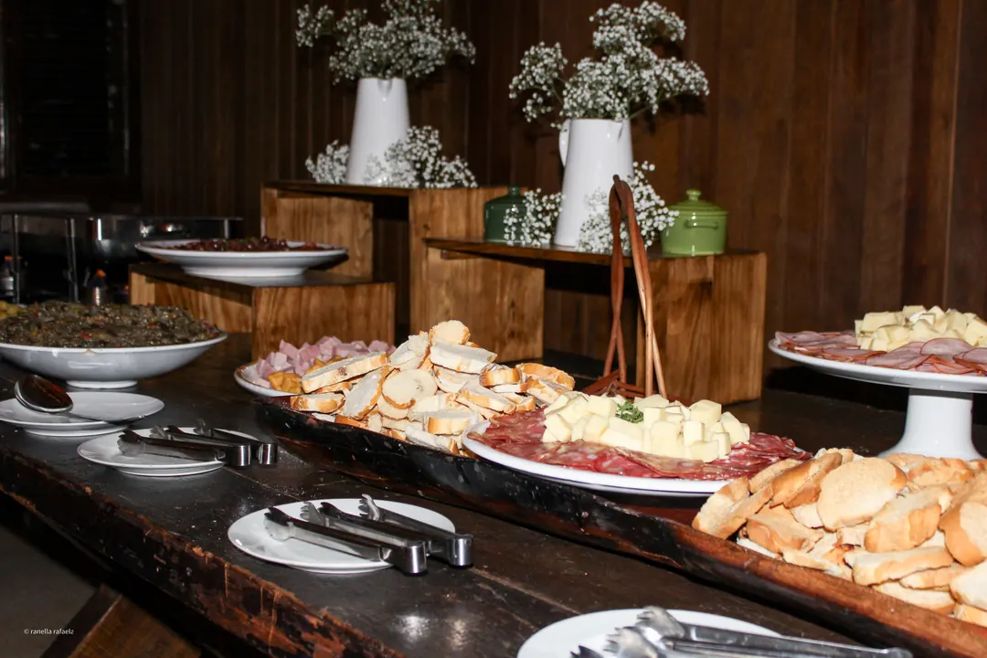Buffet de frios com queijos, pães e salames sobre uma mesa rústica, decorada com flores brancas e folhagens.