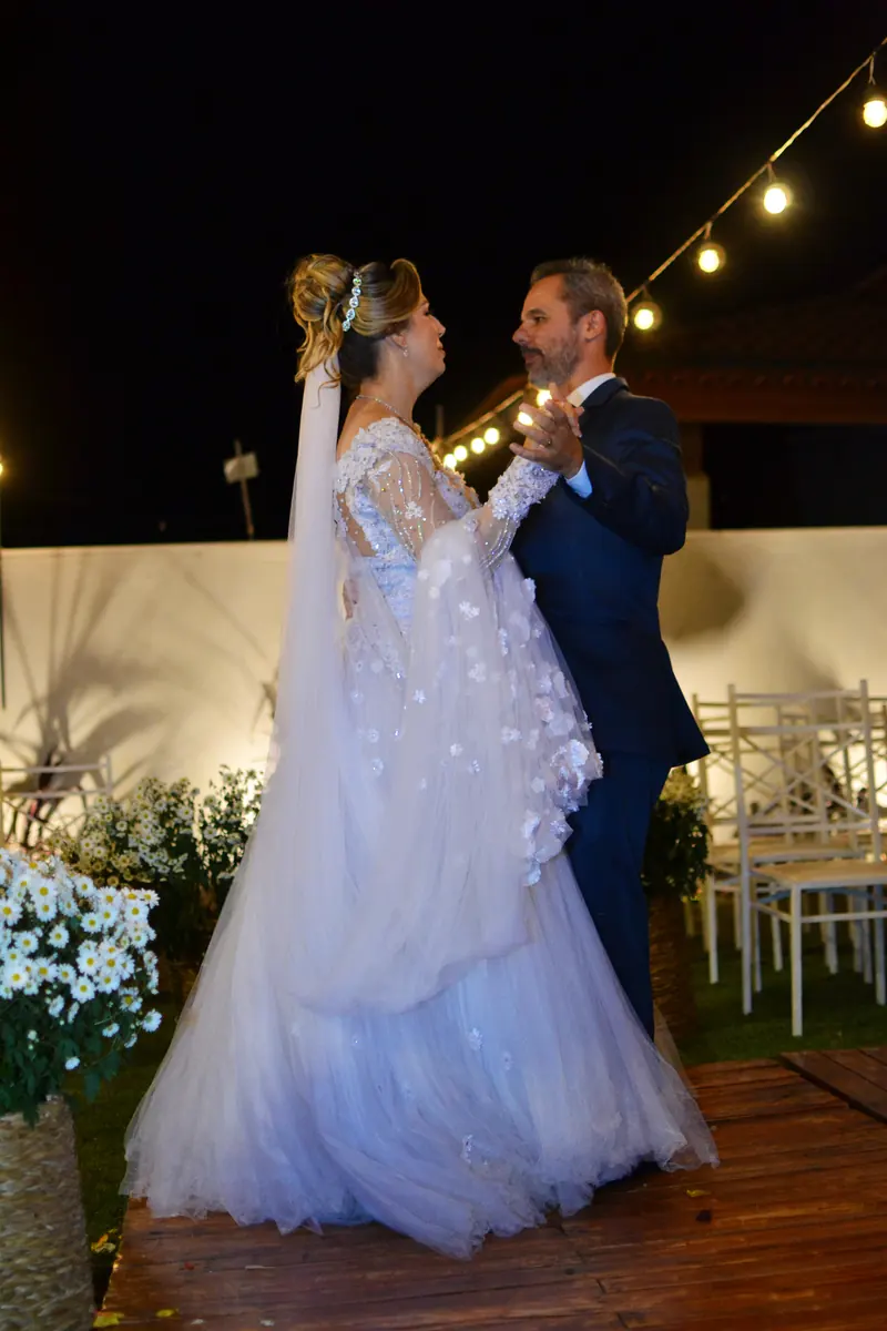 Shirley e Tiago juntos no caminho de madeira da cerimônia à noite, sob cordões de luzes e entre arranjos de flores brancas.