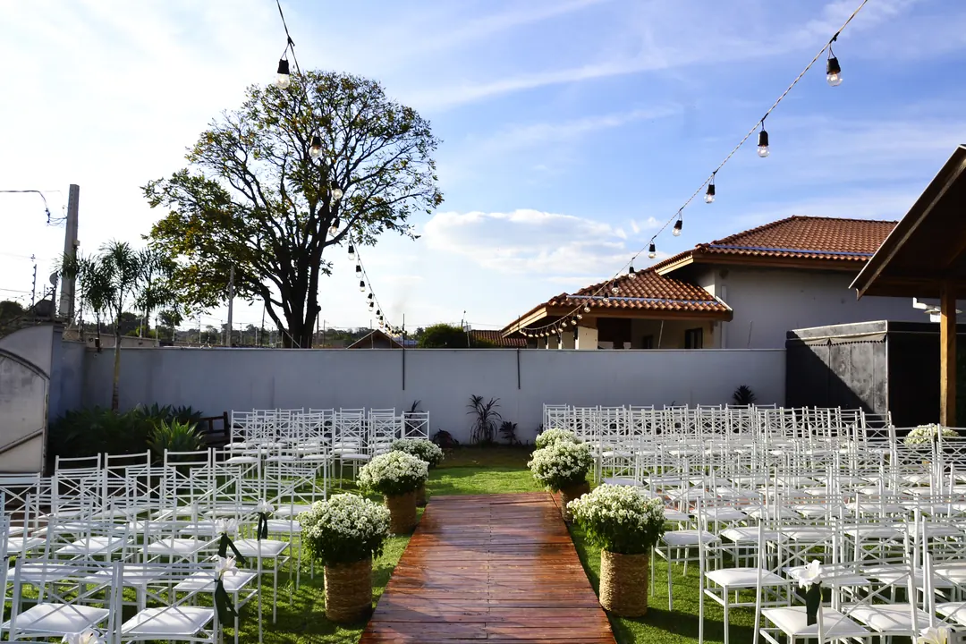 Cenário da cerimônia de Shirley e Tiago ao ar livre, com caminho de madeira, cadeiras brancas, flores e cordões de luzes.