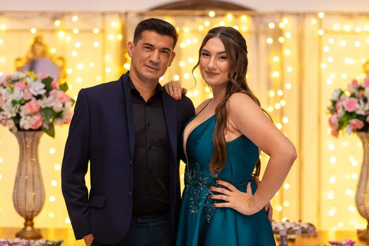 Jovem de vestido azul-petróleo ao lado de um homem de terno, diante de flores claras e luzes douradas.